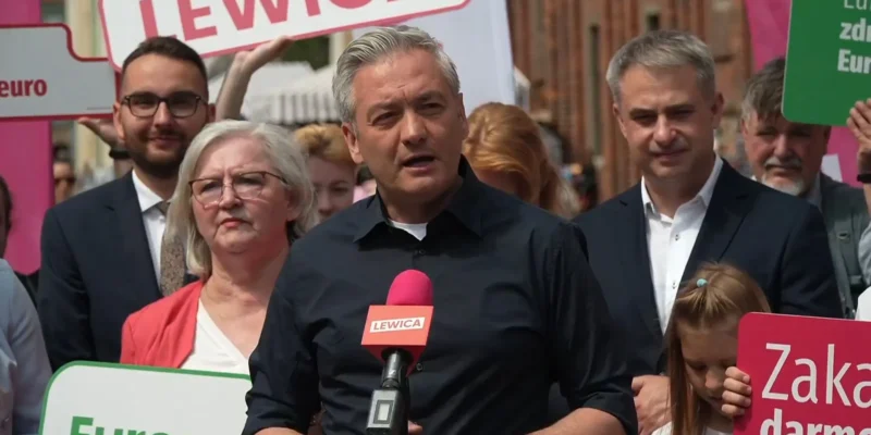 Polityk w czarnej koszuli przemawia do mikrofonu z logiem Lewica podczas zgromadzenia wyborczego, otoczony tłumem i kolorowymi plakatami.