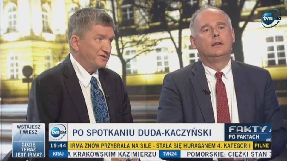 Dwóch mężczyzn w garniturach rozmawia w studiu telewizyjnym podczas programu informacyjnego, z paskiem informacyjnym na dole ekranu i logiem stacji w prawym górnym rogu.