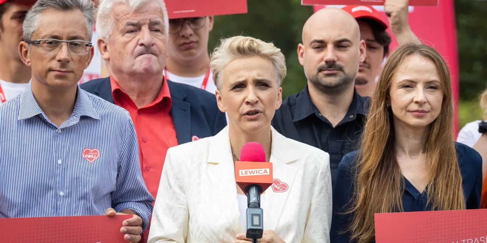 Kobieta o blond włosach w białym garniturze przemawia do mikrofonu podczas wiecu Lewicy, otoczona innymi zwolennikami trzymającymi czerwone plakaty z napisem Lewica.