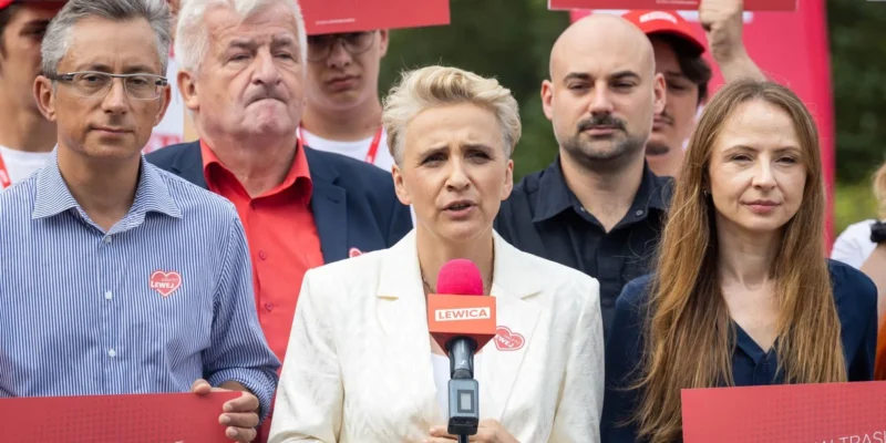 Kobieta o blond włosach w białym garniturze przemawia do mikrofonu podczas wiecu Lewicy, otoczona innymi zwolennikami trzymającymi czerwone plakaty z napisem Lewica.