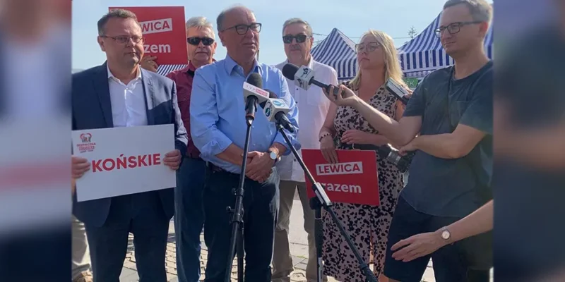 Na zdjęciu widoczna jest grupa osób podczas konferencji prasowej, trzymających transparenty z hasłami związanymi z polityką, w słonecznym otoczeniu.