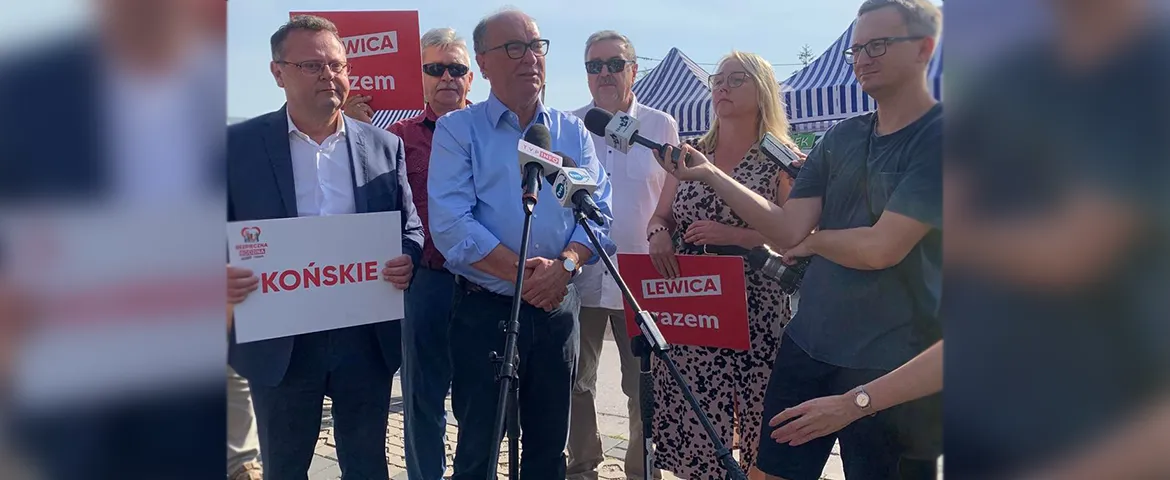 Na zdjęciu widoczna jest grupa osób podczas konferencji prasowej, trzymających transparenty z hasłami związanymi z polityką, w słonecznym otoczeniu.