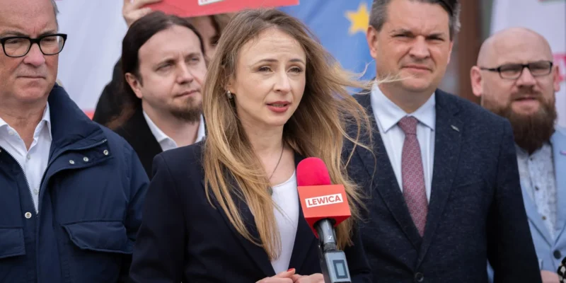 Kobieta przemawia do mikrofonu z logiem Lewicy podczas konferencji prasowej, otoczona przez grupę mężczyzn.