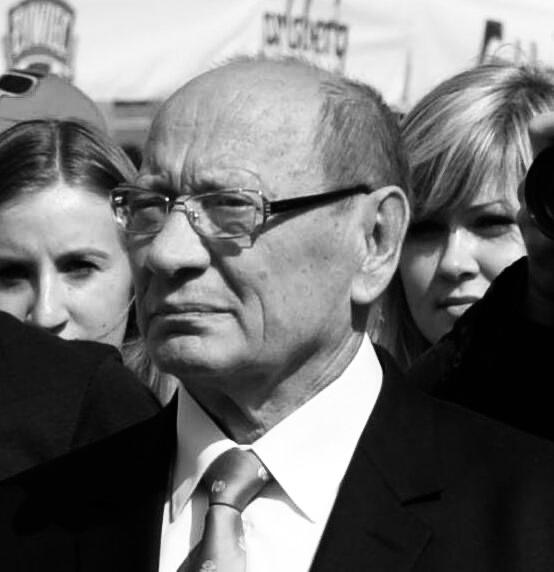 Mężczyzna w garniturze i okularach stoi wśród tłumu na czarno-białej fotografii.