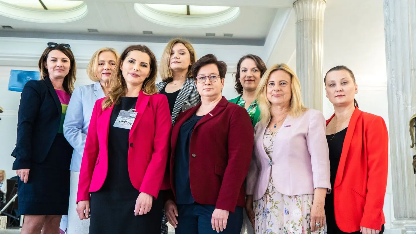 Na zdjęciu znajduje się grupa dziewięciu kobiet stojących razem w jasnym wnętrzu, ubrane w eleganckie stroje, prezentujące różne style i kolory.