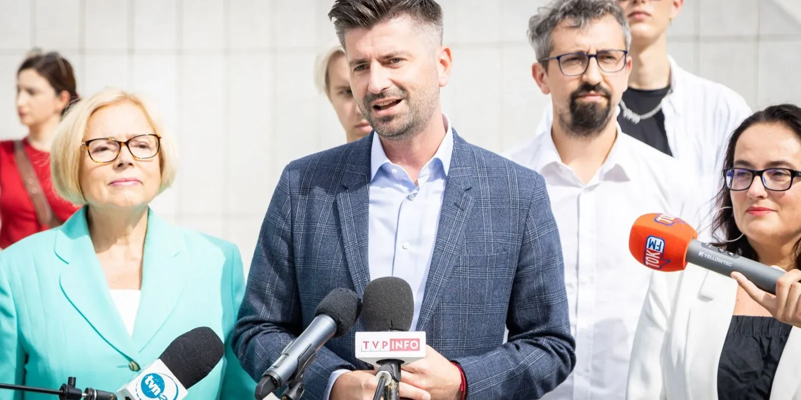 Mężczyzna w granatowym garniturze wygłasza oświadczenie na konferencji prasowej, otoczony dziennikarzami z mikrofonami.