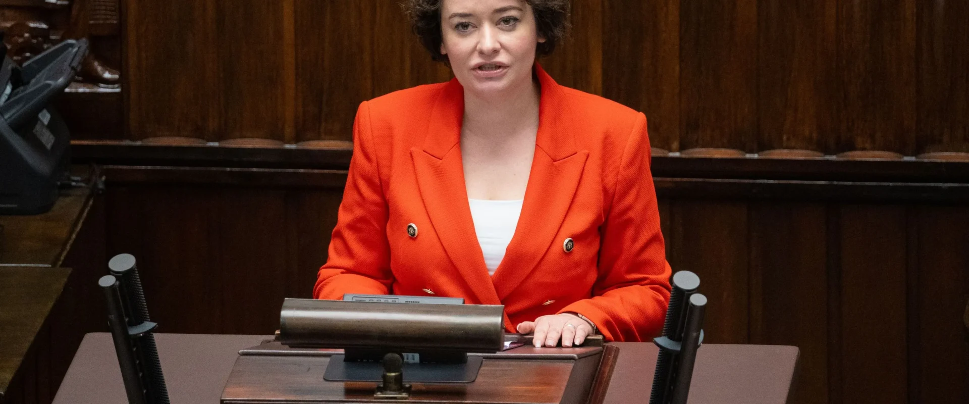 Kobieta w pomarańczowym garniturze przemawia z mównicy w sejmie.