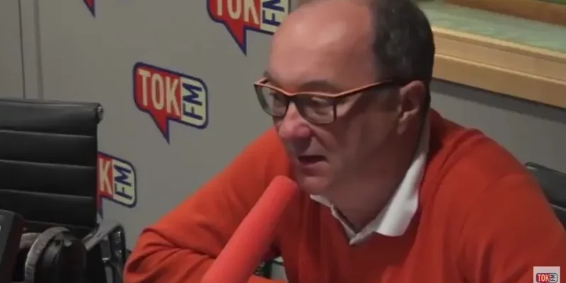 Mężczyzna w pomarańczowym swetrze i okularach mówi do mikrofonu w studiu radiowym z widocznym logiem TOK FM.