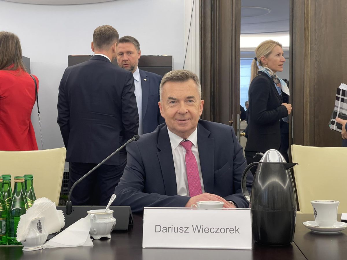 Mężczyzna w garniturze siedzi przy konferencyjnym stole z mikrofonem, dzbankiem kawowym i filiżankami; w tle widoczni inni uczestnicy spotkania.