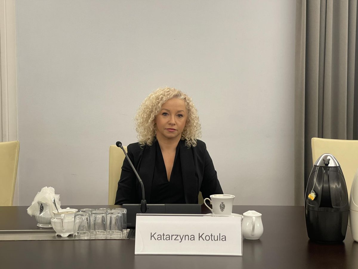 Kobieta o kręconych blond włosach siedzi przy czarnym stole w sali konferencyjnej, przed sobą ma mikrofon oraz białą filiżankę.