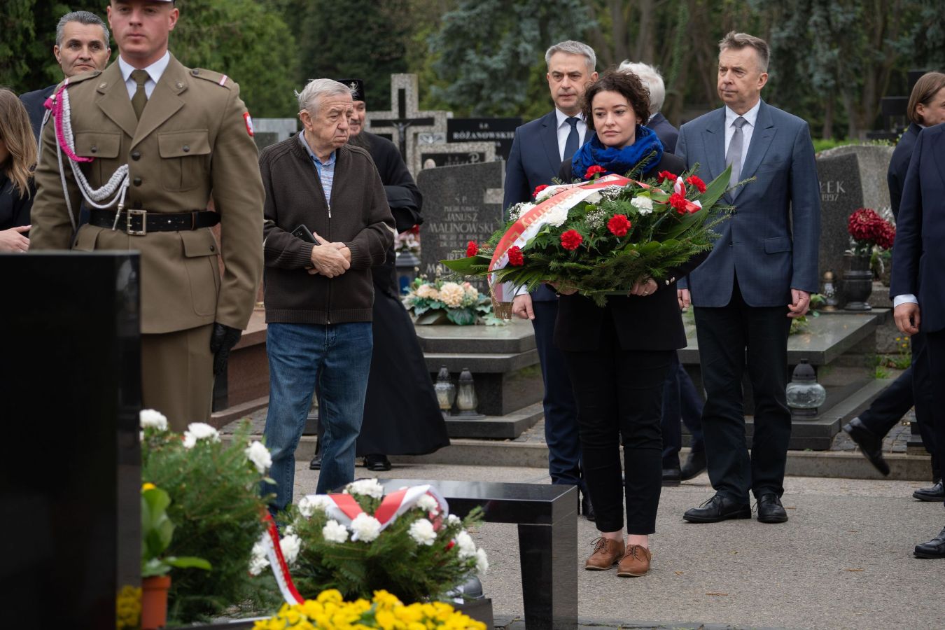 Grupa osób na cmentarzu podczas 14. rocznicy katastrofy smoleńskiej, kobieta trzyma duży wieniec z czerwonych i białych kwiatów.