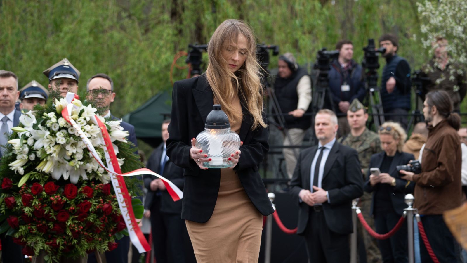 Uczestniczka uroczystości upamiętniającej katastrofę smoleńską trzyma w dłoniach szklany lampion, stojąc obok dużego kwiatowego wieńca i otoczonej kamerami grupy mediów.