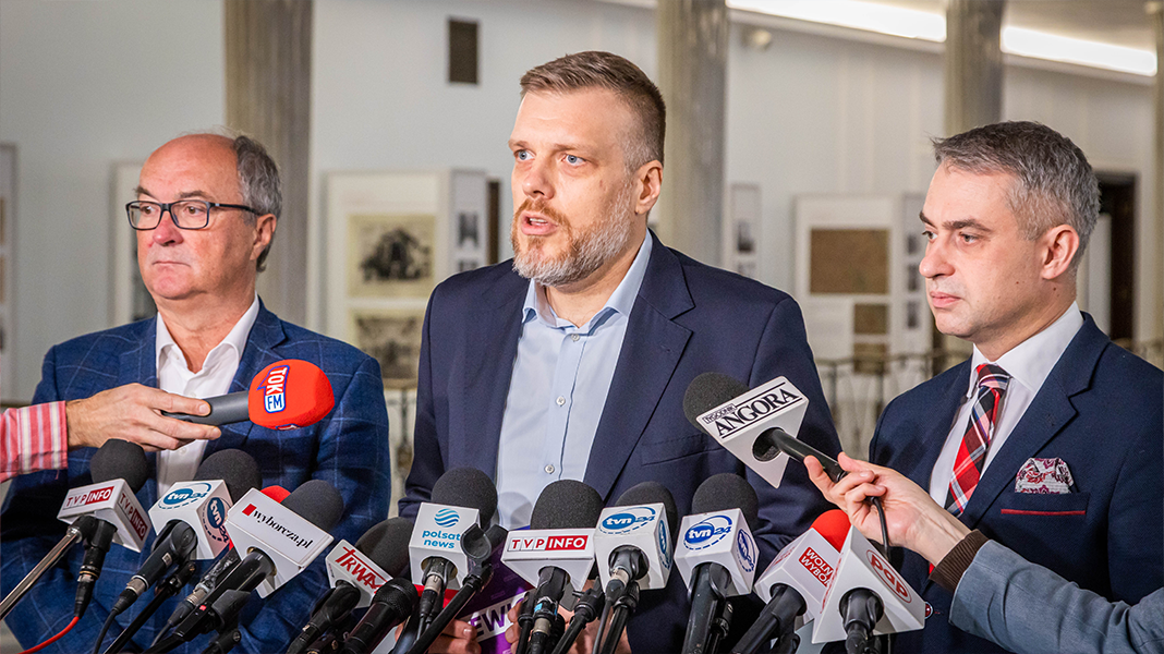 Na zdjęciu widoczni są trzej mężczyźni podczas konferencji prasowej, otoczeni mikrofonami mediów.