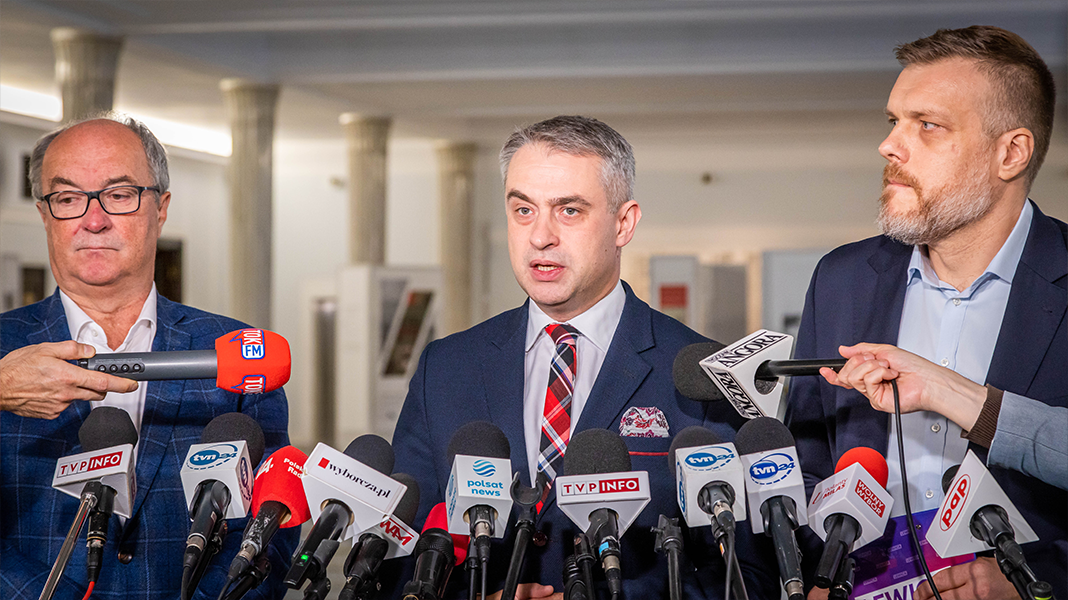 Na zdjęciu trzech mężczyzn stoi przed mikrofonami i aparatem reporterskim, podczas konferencji prasowej.