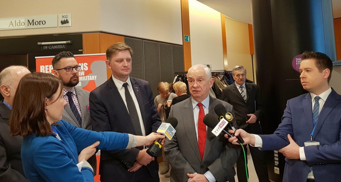 Mężczyzna w garniturze i czerwonym krawacie udziela wywiadu, otoczony dziennikarzami z mikrofonami i kilkoma osobami w tle.