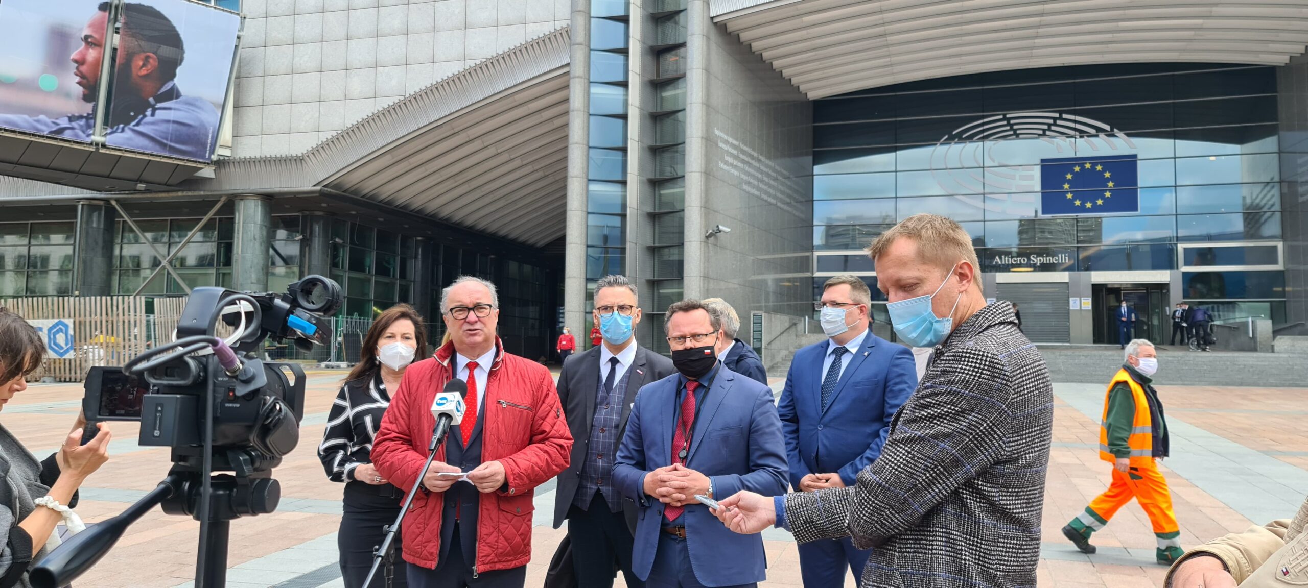 Grupa osób w garniturach i reporterzy prowadzą konferencję prasową na placu przed wejściem do budynku Parlamentu Europejskiego, z flagą Unii Europejskiej widoczną w tle.