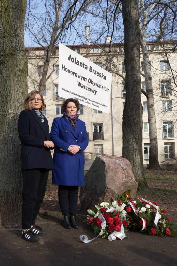 Dwie kobiety stoją w parku obok banneru upamiętniającego osobę, a na ziemi leżą kwiaty i wieńce, z budynkiem w tle.