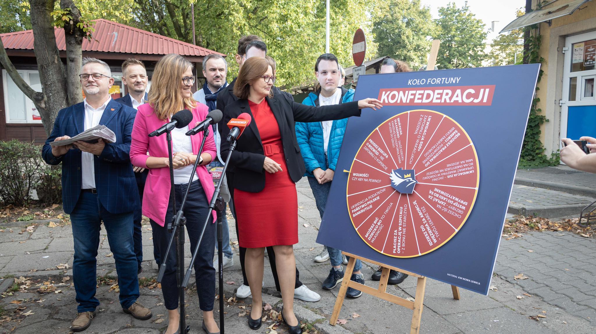 Grupa ludzi na zewnątrz przy dużym kole fortuny Konfederacji ustawionym na sztaludze, kobieta w czerwonej sukience wskazuje koło podczas konferencji prasowej.