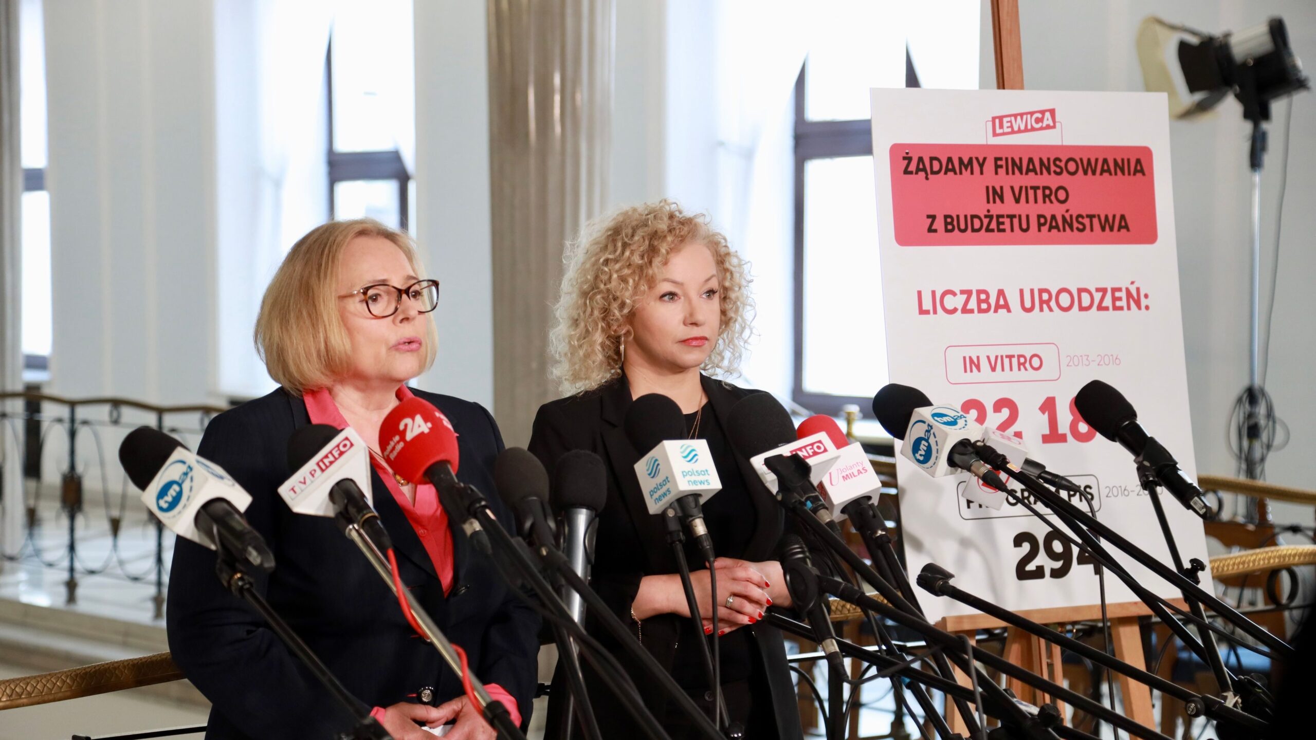 Dwie kobiety stoją przy mikrofonach podczas konferencji prasowej, obok plakatu z napisem „Żądamy finansowania IN VITRO z budżetu państwa”.
