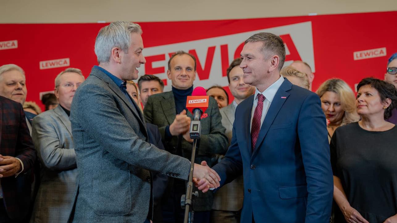 Dwóch mężczyzn w garniturach ściska sobie dłonie podczas konferencji prasowej Lewicy, na tle czerwonego tła z logotypem Lewicy i zgromadzonych zwolenników.