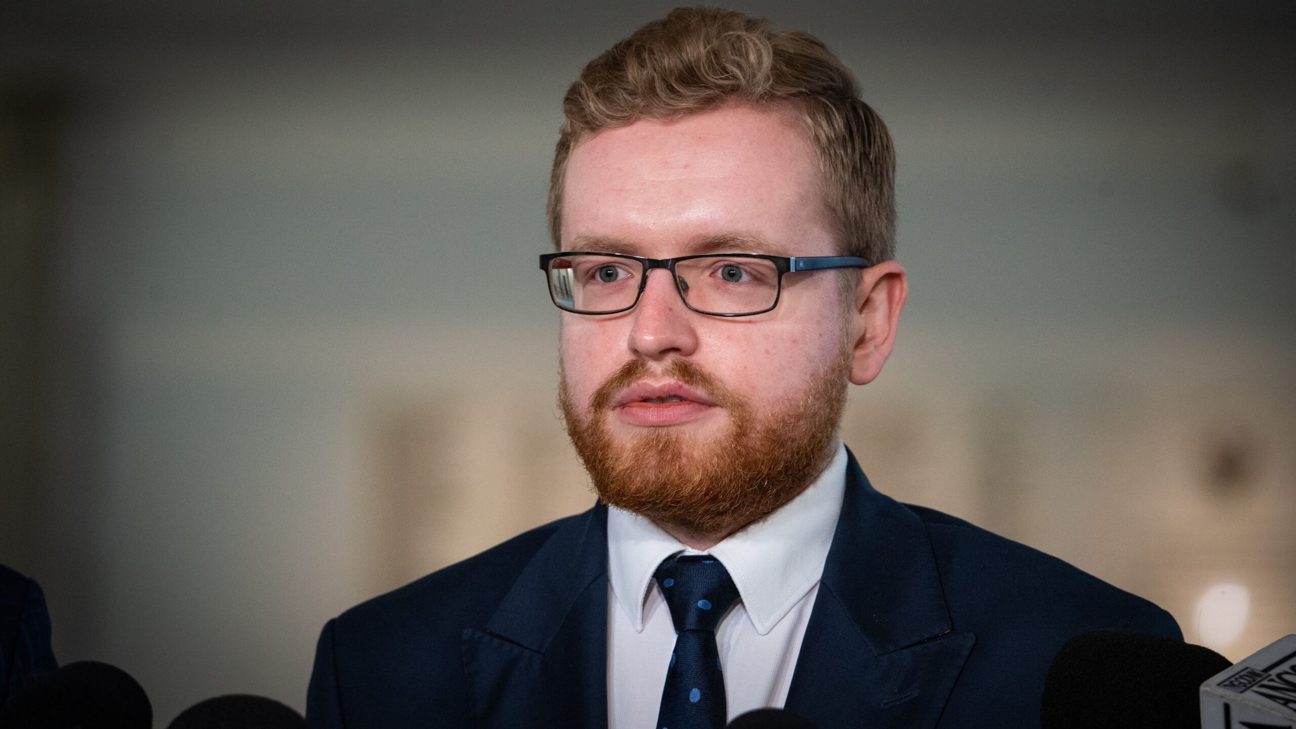 Mężczyzna w garniturze i okularach przemawia do mikrofonów podczas konferencji prasowej.