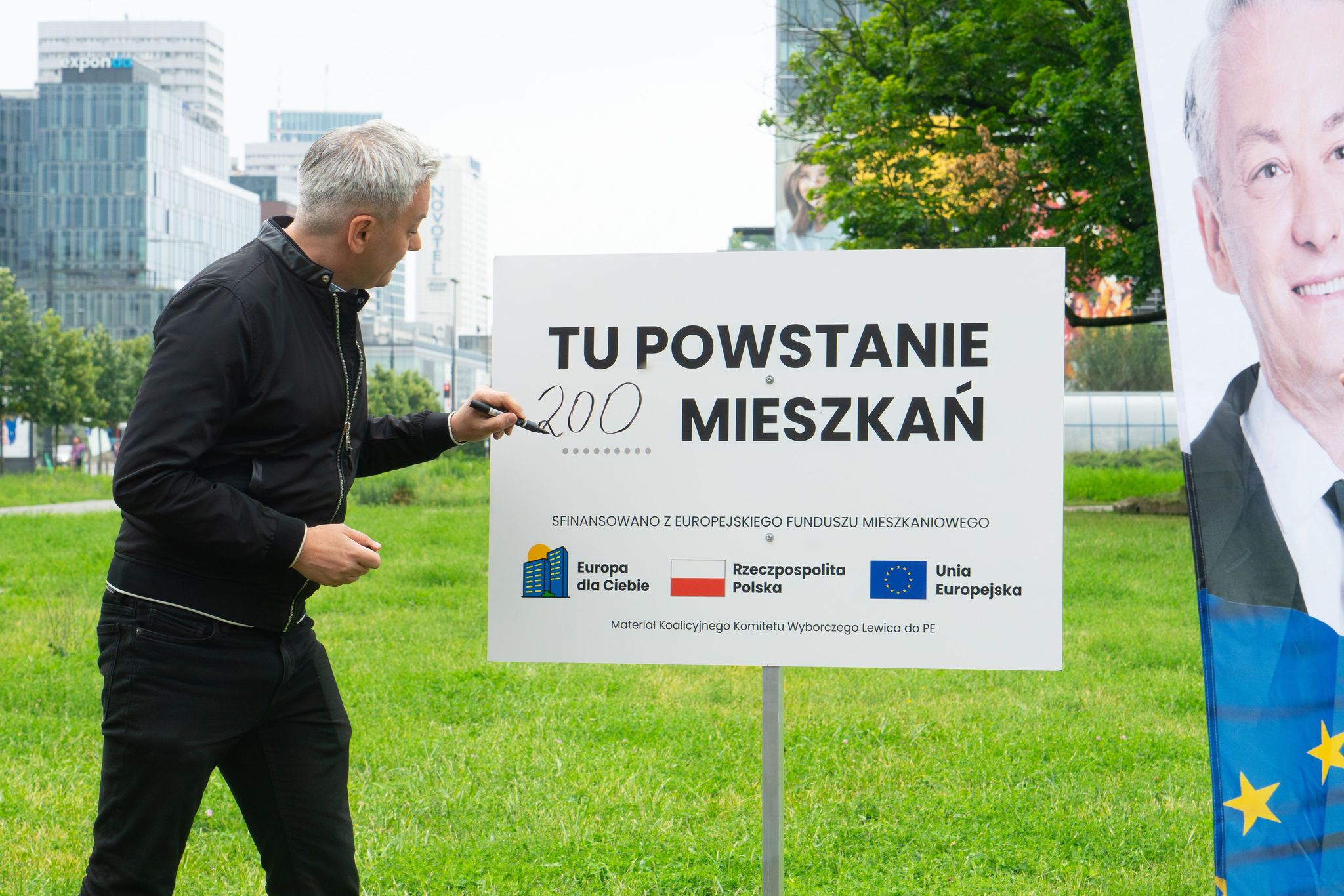 Mężczyzna w czarnej kurtce podpisuje tablicę informującą o powstaniu 200 mieszkań, ustawioną na zielonym skwerze z logotypami funduszy unijnych na dole.