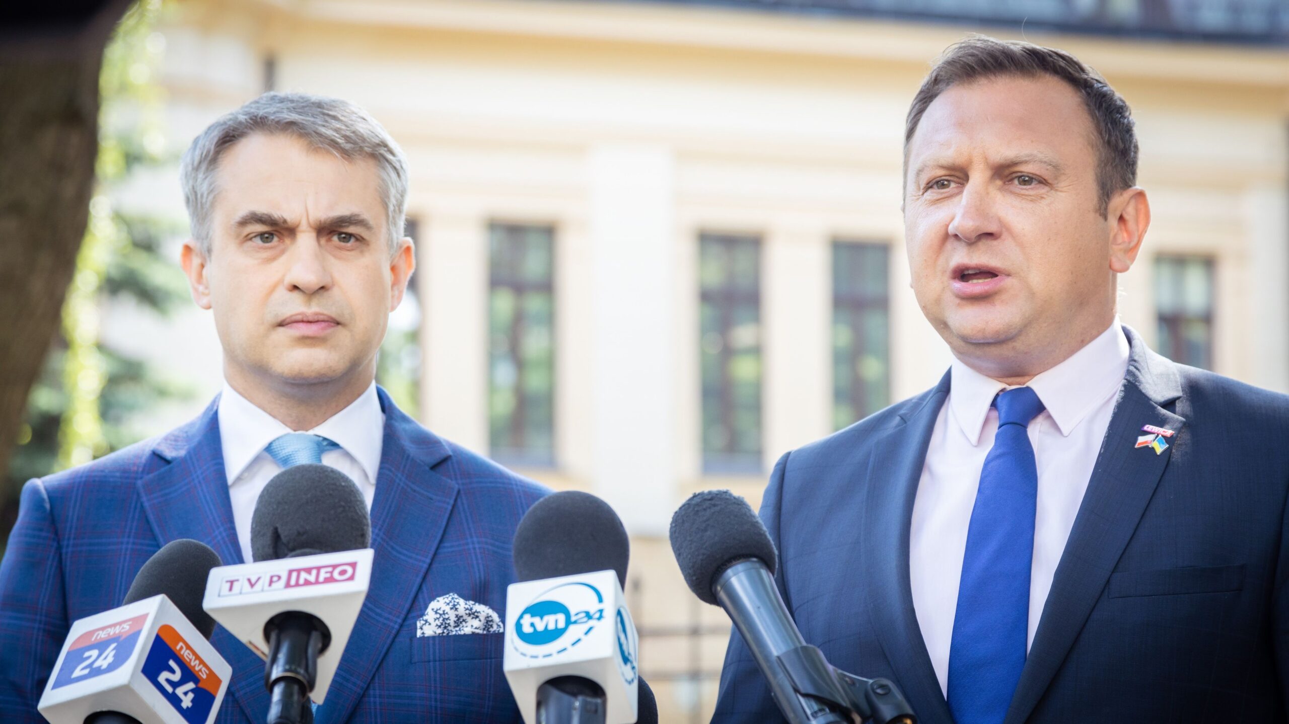 Dwóch mężczyzn w garniturach podczas konferencji prasowej stojących przy mikrofonach przed budynkiem.