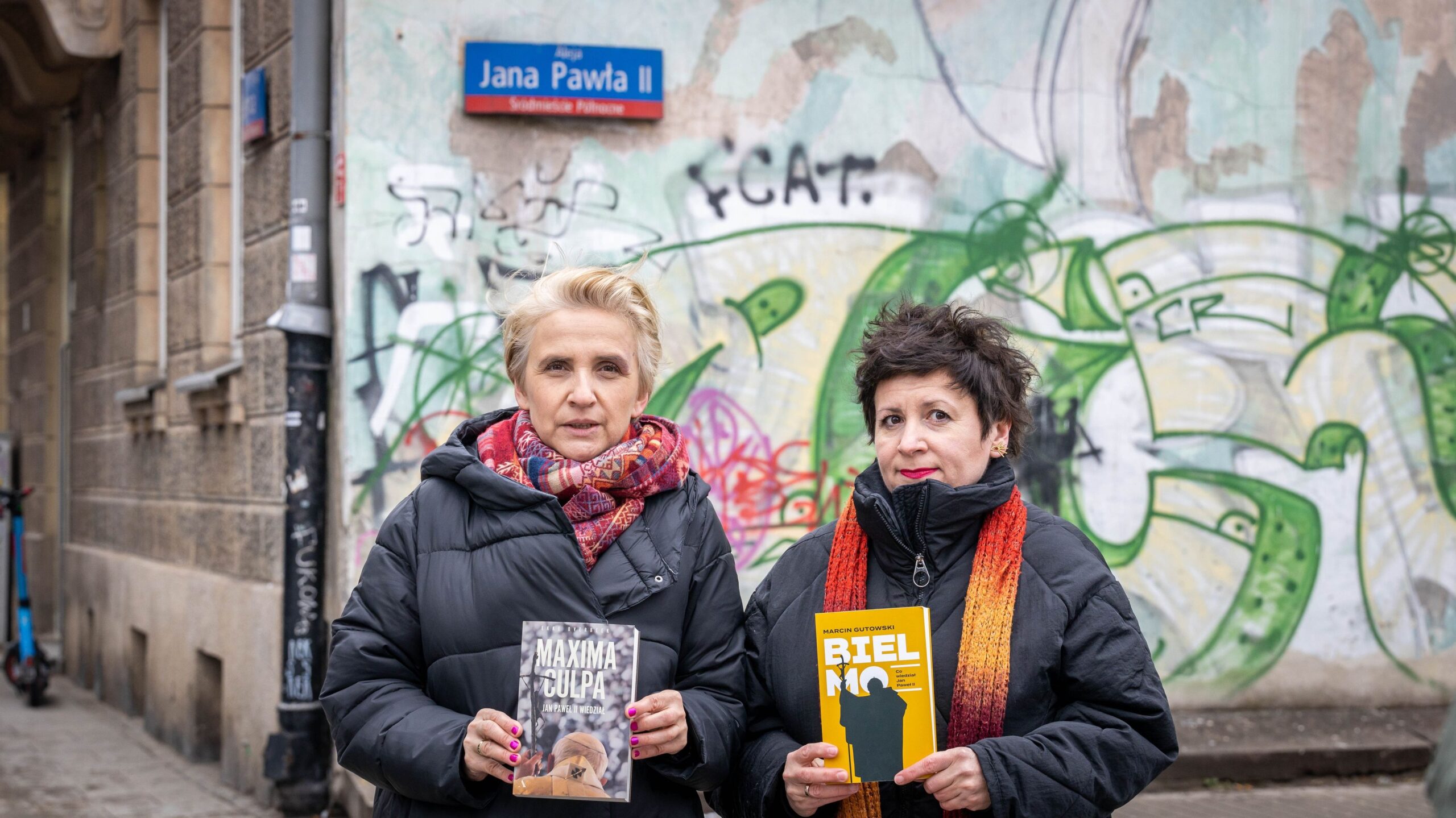 Dwie kobiety trzymające książki stoją przed ścianą z graffiti, obok znaku ulicy Jana Pawła II.