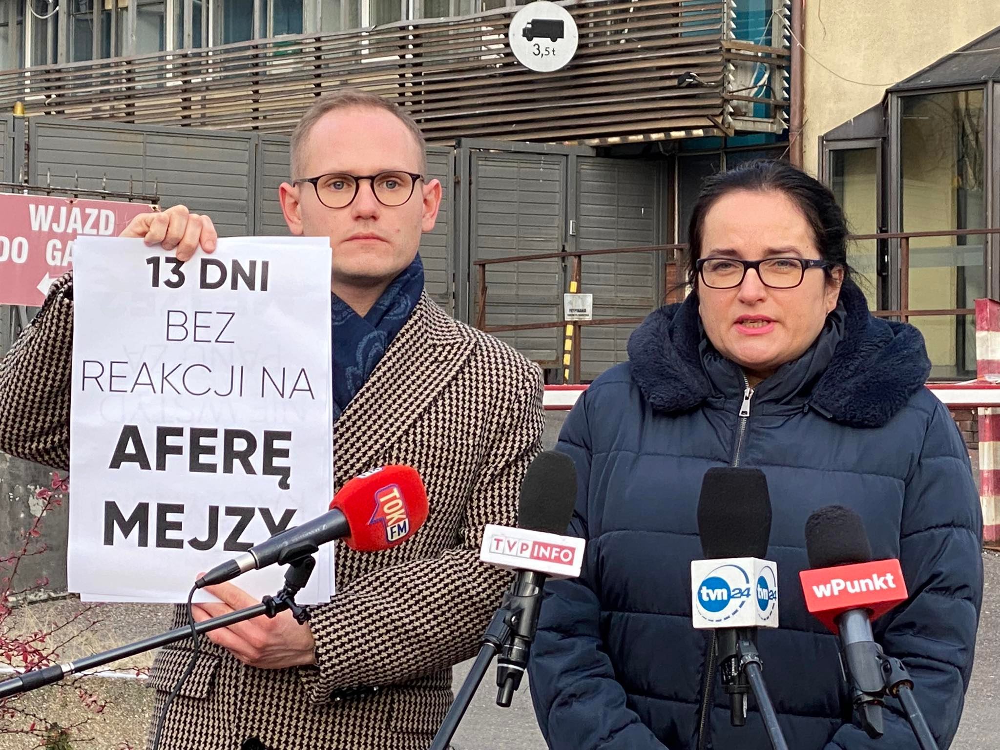 Dwie osoby stoją przy mikrofonach podczas konferencji prasowej Lewicy; mężczyzna po lewej trzyma plakat z napisem „13 DNI BEZ REAKCJI NA AFERĘ MEJZY”.