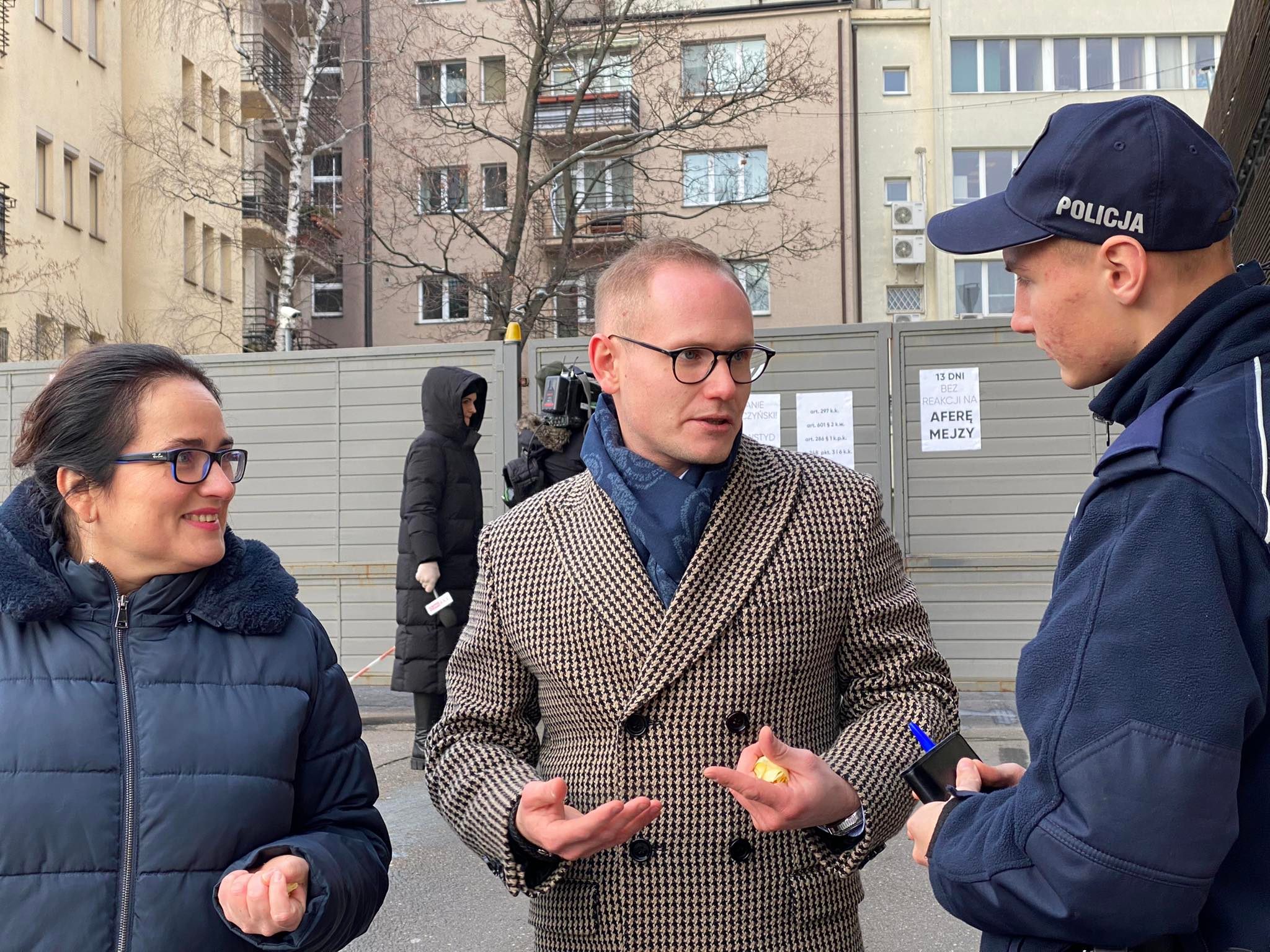 Na zdjęciu widoczna jest rozmowa między dwoma osobami, z których jedna jest policjantem, a w tle widać budynki mieszkalne i osobę w czarnym płaszczu.