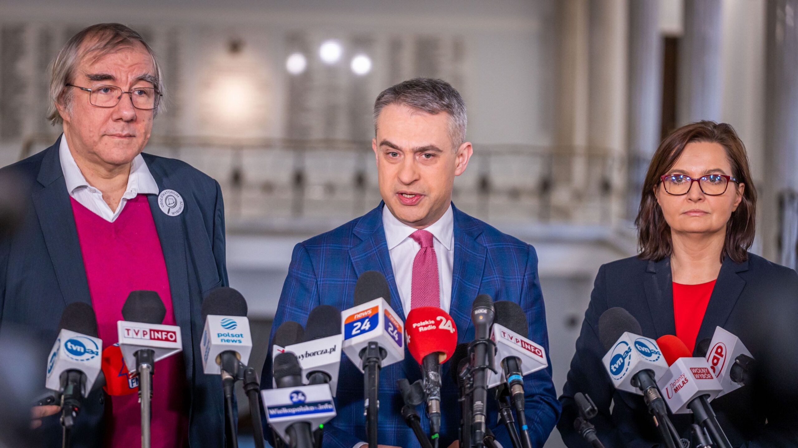 Trzech polityków w garniturach stoi przy wielu mikrofonach podczas konferencji prasowej o Kodeksie Wyborczym.