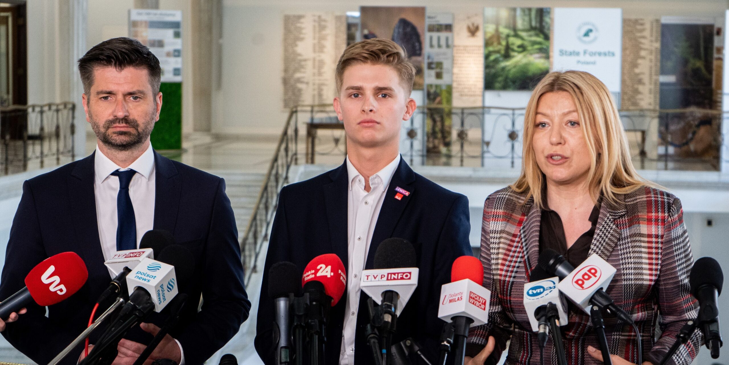 Trzech mówców stoi przed dziennikarzami, otoczeni mikrofonami, w przestronnej sali przypominającej miejsce konferencji prasowej.