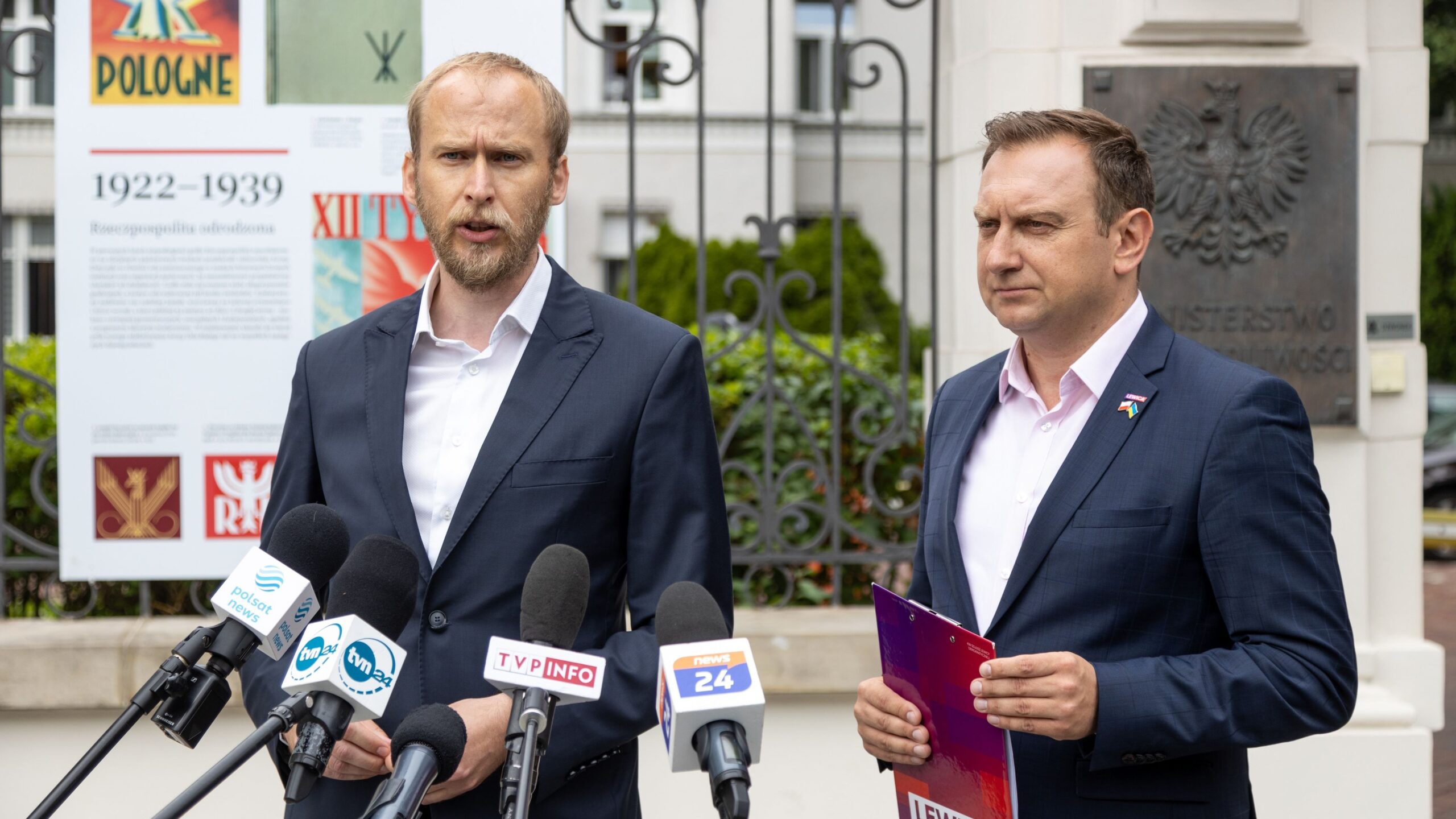 Dwóch mężczyzn w garniturach udziela konferencji prasowej przed zestawem mikrofonów, z tablicą informacyjną i metalową bramą w tle.