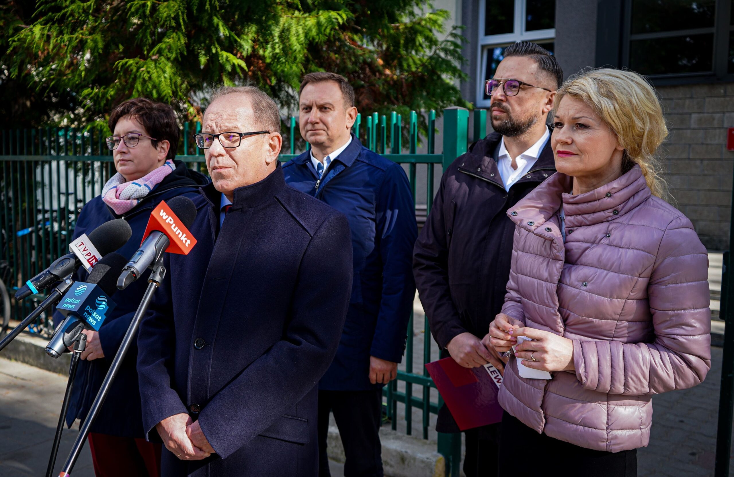 Na zdjęciu widać grupę pięciu osób stojących przed mikrofonami podczas konferencji prasowej na świeżym powietrzu.