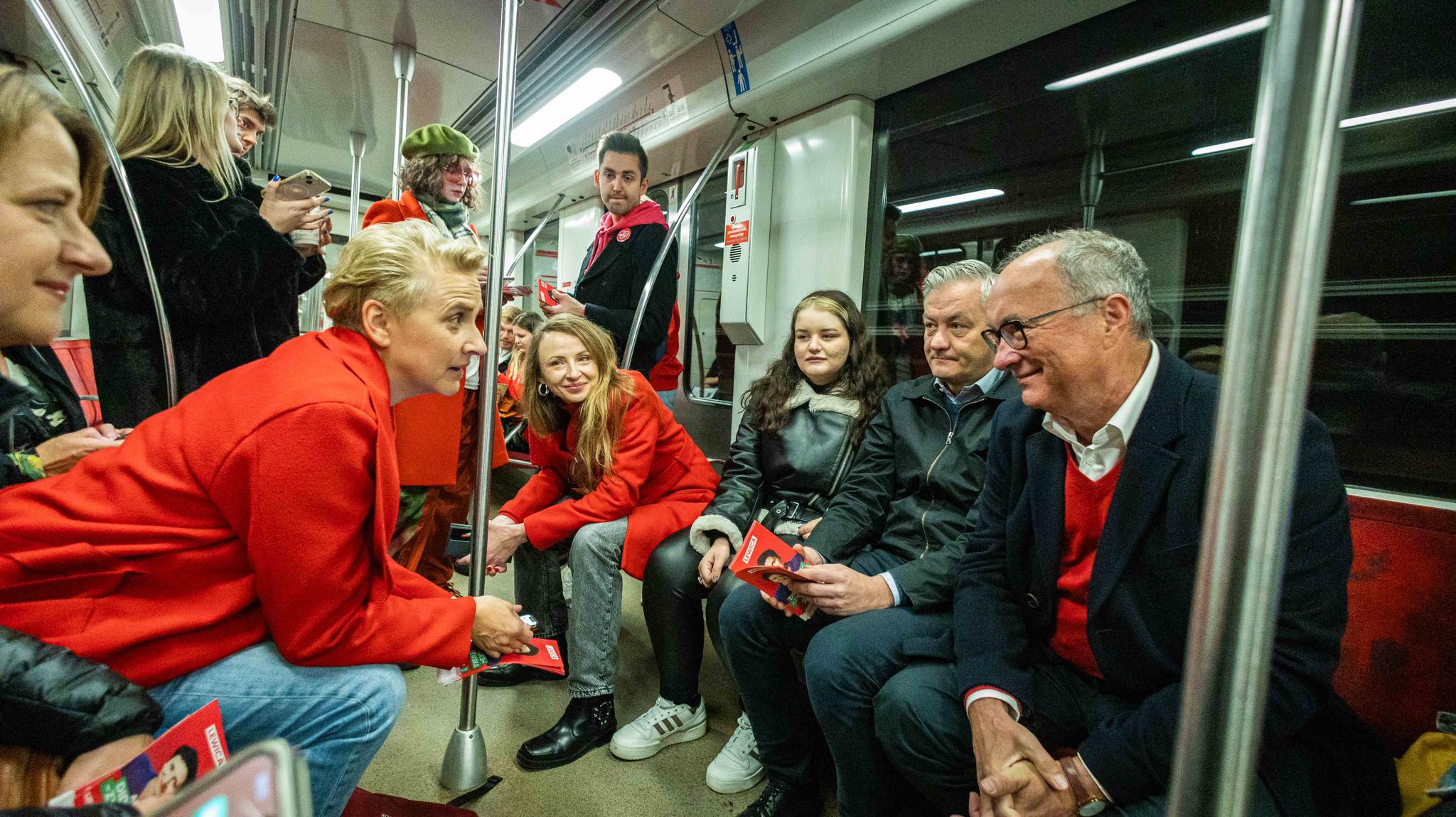 Grupa ludzi w czerwonych ubraniach siedzi i stoi w wagonie metra, część z nich rozdaje ulotki podczas wydarzenia o charakterze politycznym.