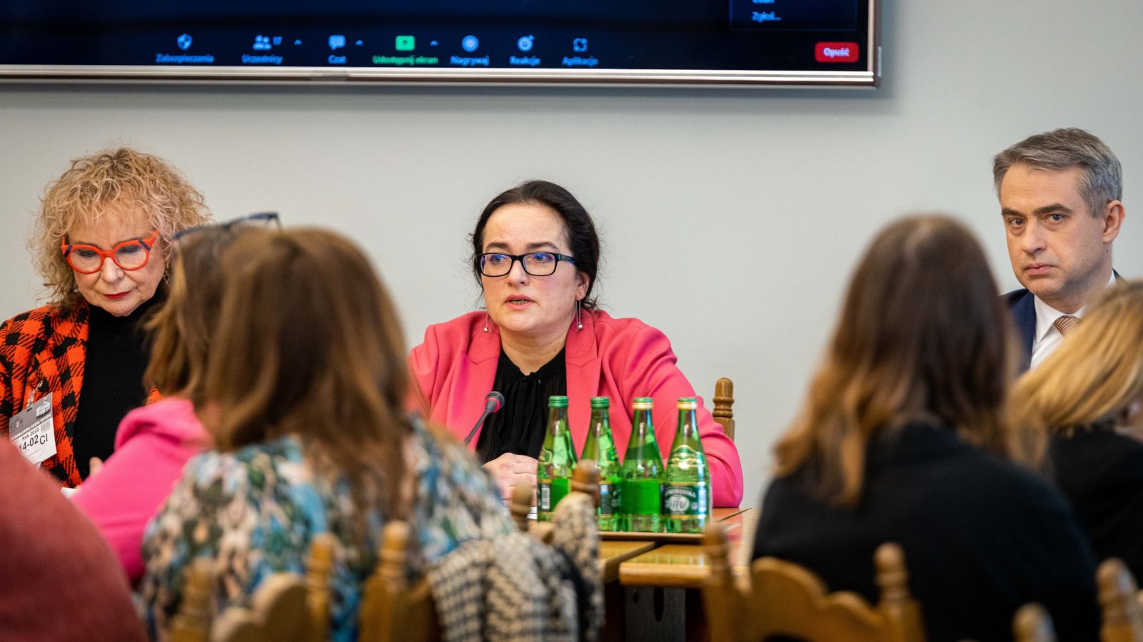 Na zdjęciu przedstawiona jest trójka uczestników konferencji, siedzących przy stole, a w tle widoczny jest ekran.