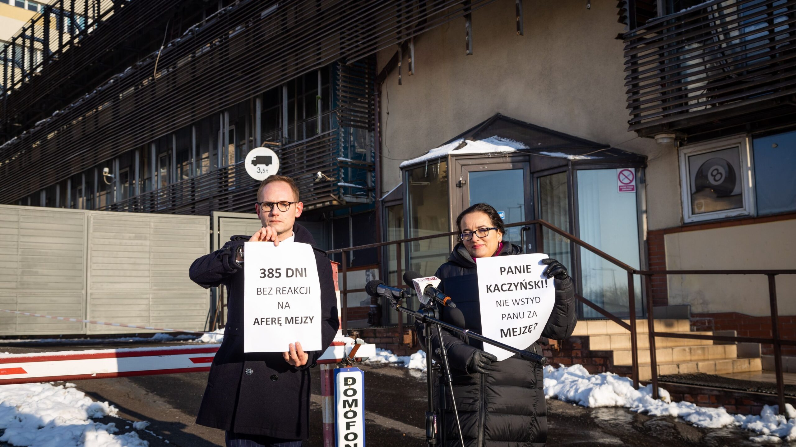 Na zdjęciu widać dwie osoby trzymające kartki z hasłami protestacyjnymi przed budynkiem, w którym leży śnieg.