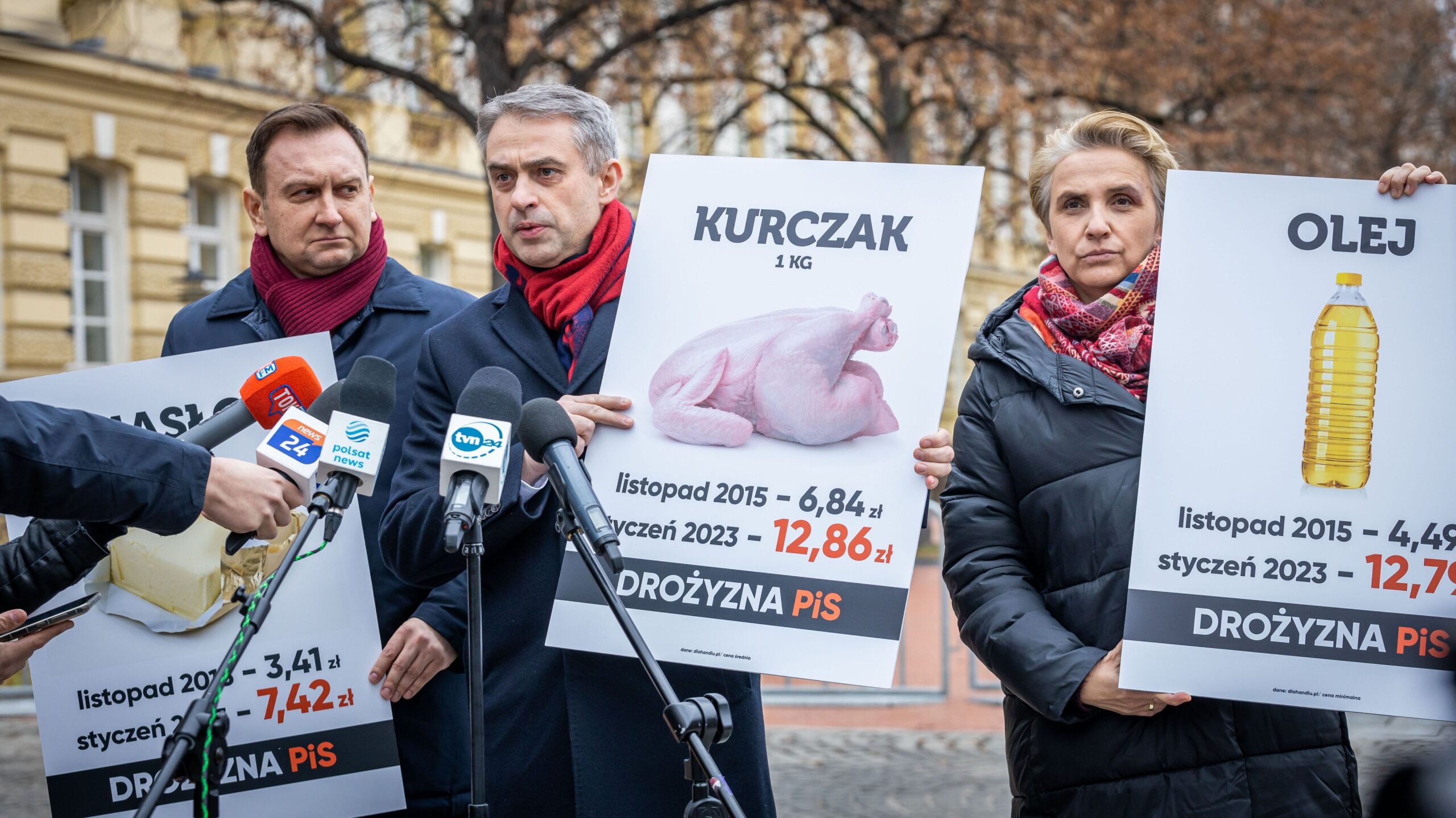 Na zdjęciu widoczna jest konferencja prasowa, w której uczestniczą trzy osoby trzymające plakaty z cenami żywności, w tym kurczaka, masła i oleju, podczas gdy mikrofony skierowane są w ich stronę.