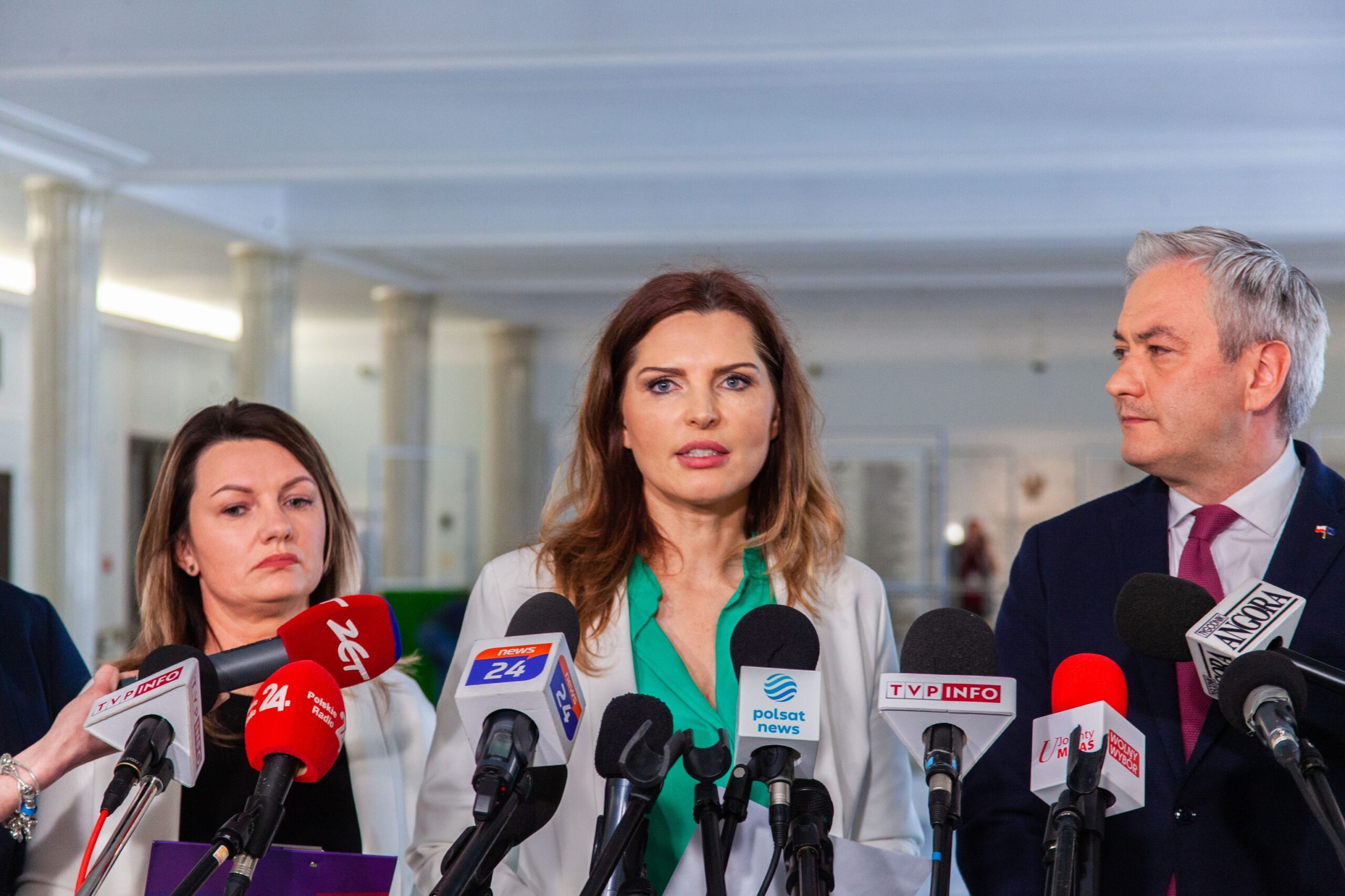 Na zdjęciu widoczni są mówcy podczas konferencji prasowej, otoczeni mikrofonami i kamerami w jasnym, przestronnym pomieszczeniu.