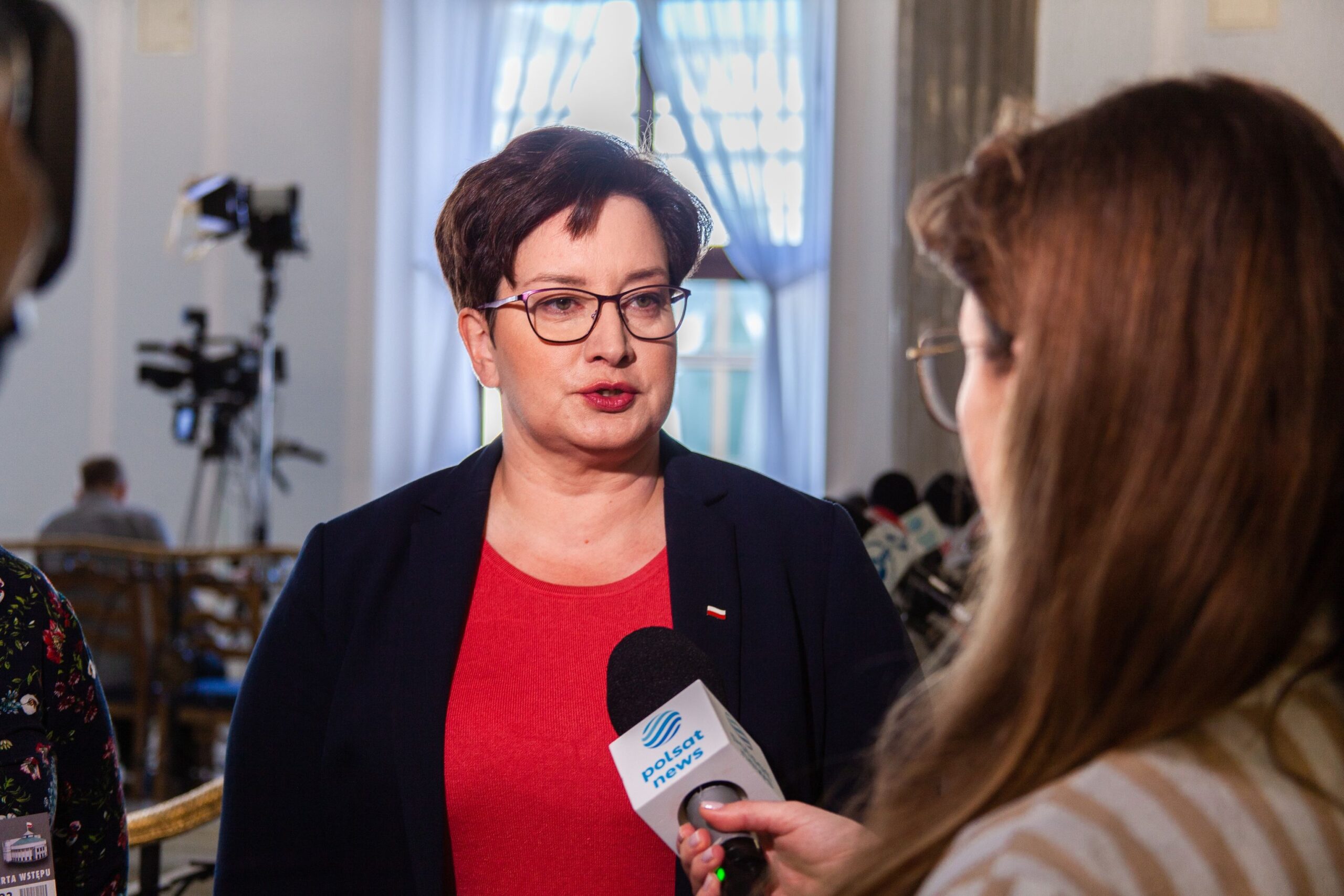 Na zdjęciu widoczna jest kobieta w okularach, ubrana w czerwoną bluzkę i granatową marynarkę, udzielająca wywiadu podczas konferencji prasowej.