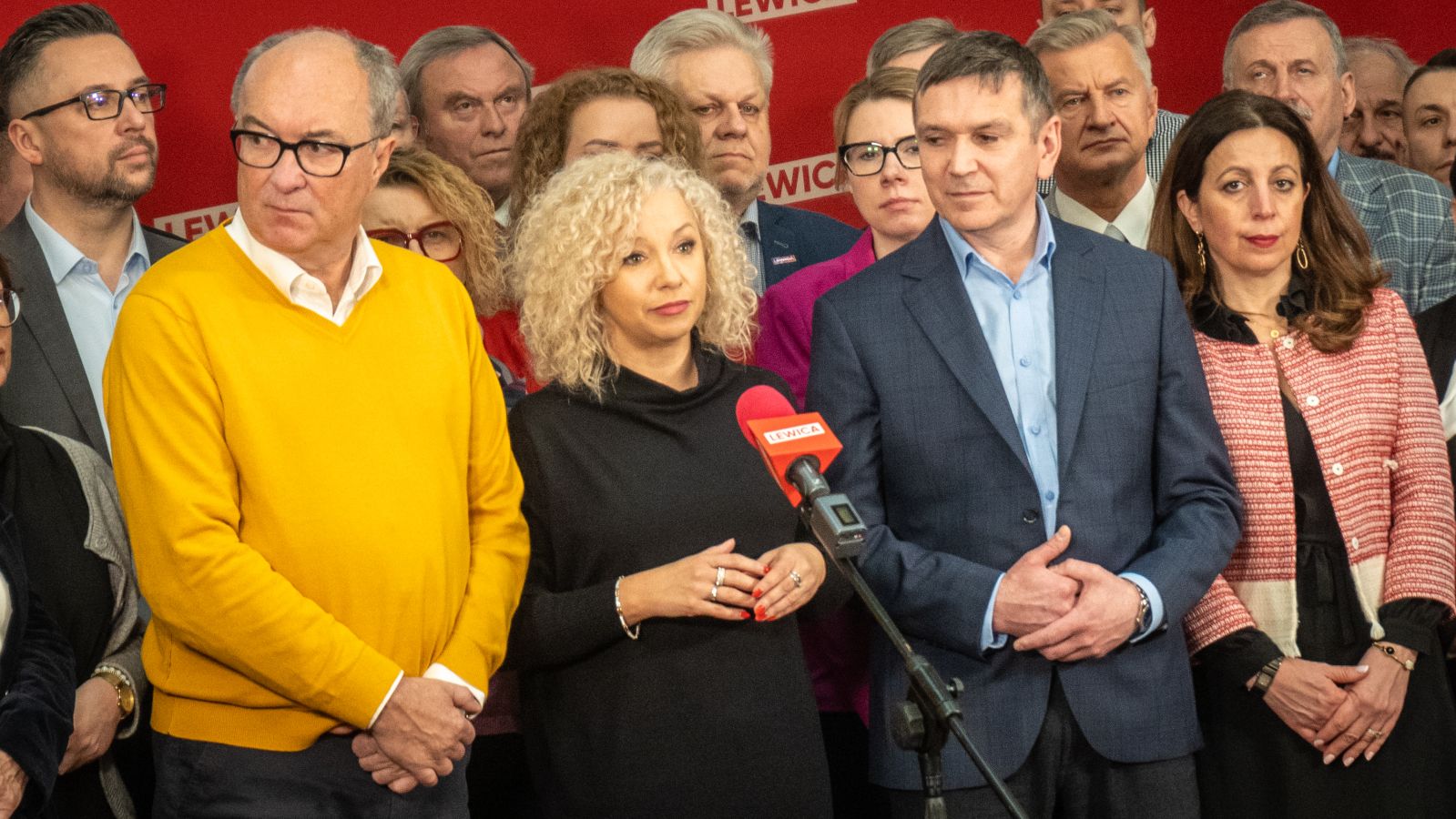 Grupa polityków Lewicy podczas konferencji prasowej, na pierwszym planie kobieta z blond lokami w czarnej sukience mówiąca do mikrofonu, otoczona innymi działaczami na czerwonym tle z logiem Lewicy.