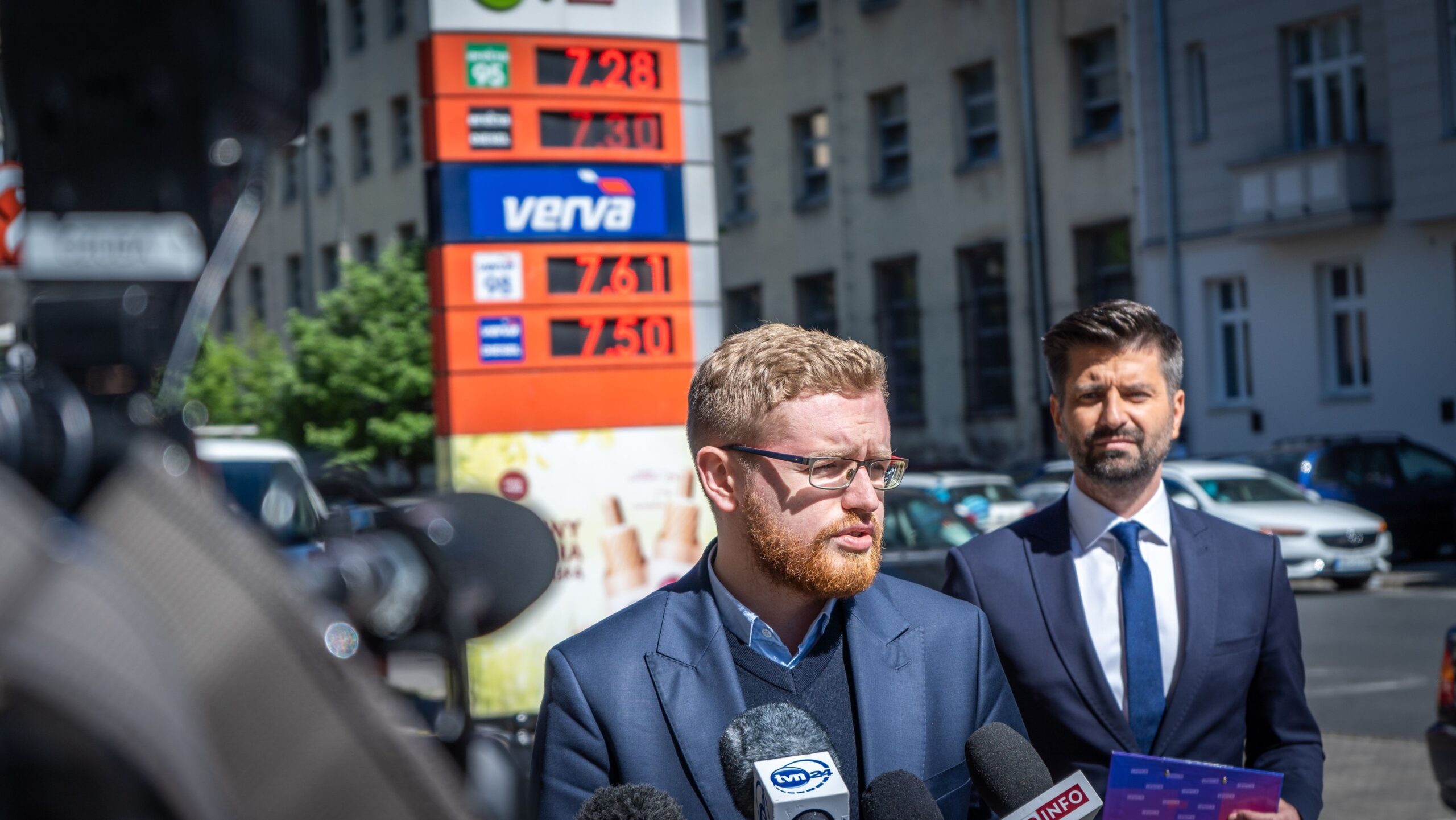 Na zdjęciu widać mężczyznę w garniturze, który przemawia do mediów w pobliżu stacji paliw, a w tle widoczne są ceny paliw.