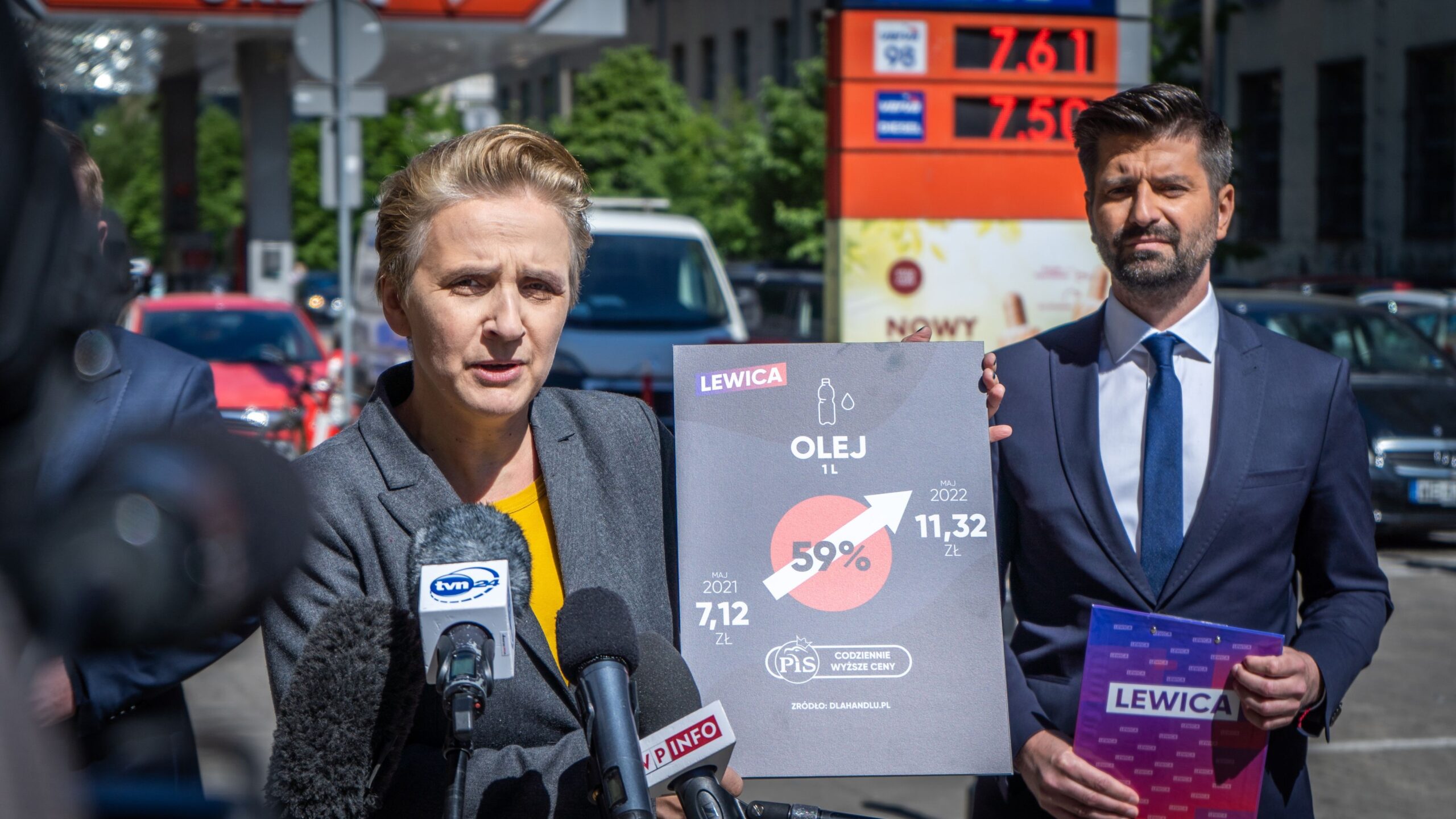 Kobieta przemawia do mediów podczas konferencji prasowej, stojąc obok mężczyzny trzymającego plakat Lewica z informacjami o cenie oleju za 1 litr, na tle stacji benzynowej.