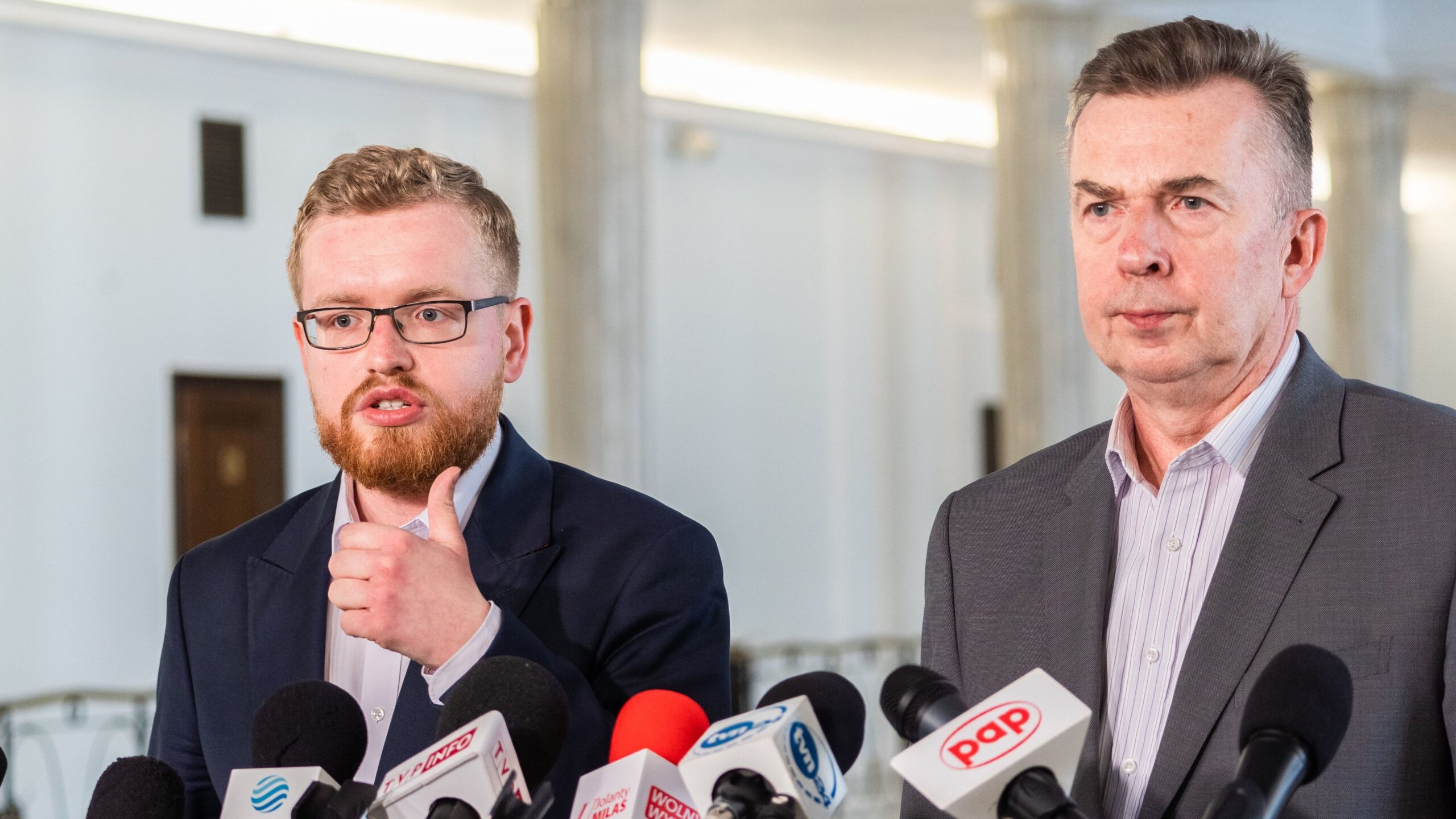 Dwa mężczyźni w garniturach stoją przed mikrofonami podczas konferencji prasowej.