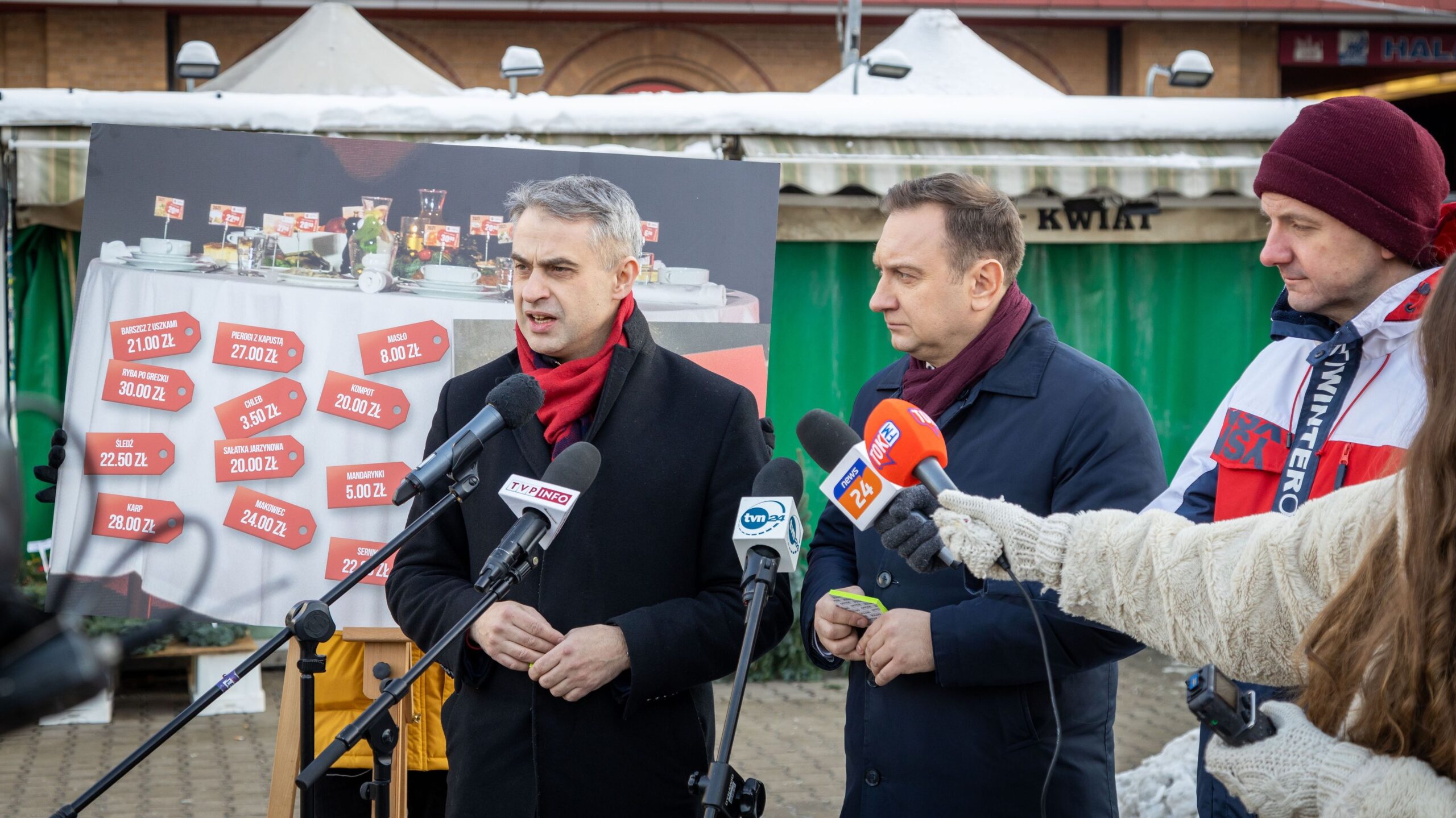 Na zdjęciu widoczna jest konferencja prasowa, podczas której trzech mężczyzn przedstawia ceny potraw wigilijnych, prezentowanych na plakacie w tle.