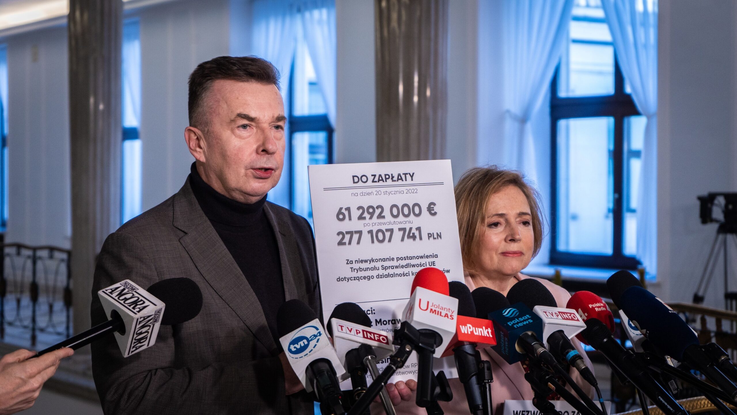 Na zdjęciu dwie osoby z branży publicznej występują na konferencji prasowej, prezentując informację o wymaganej płatności w wysokości ponad 61 milionów euro.