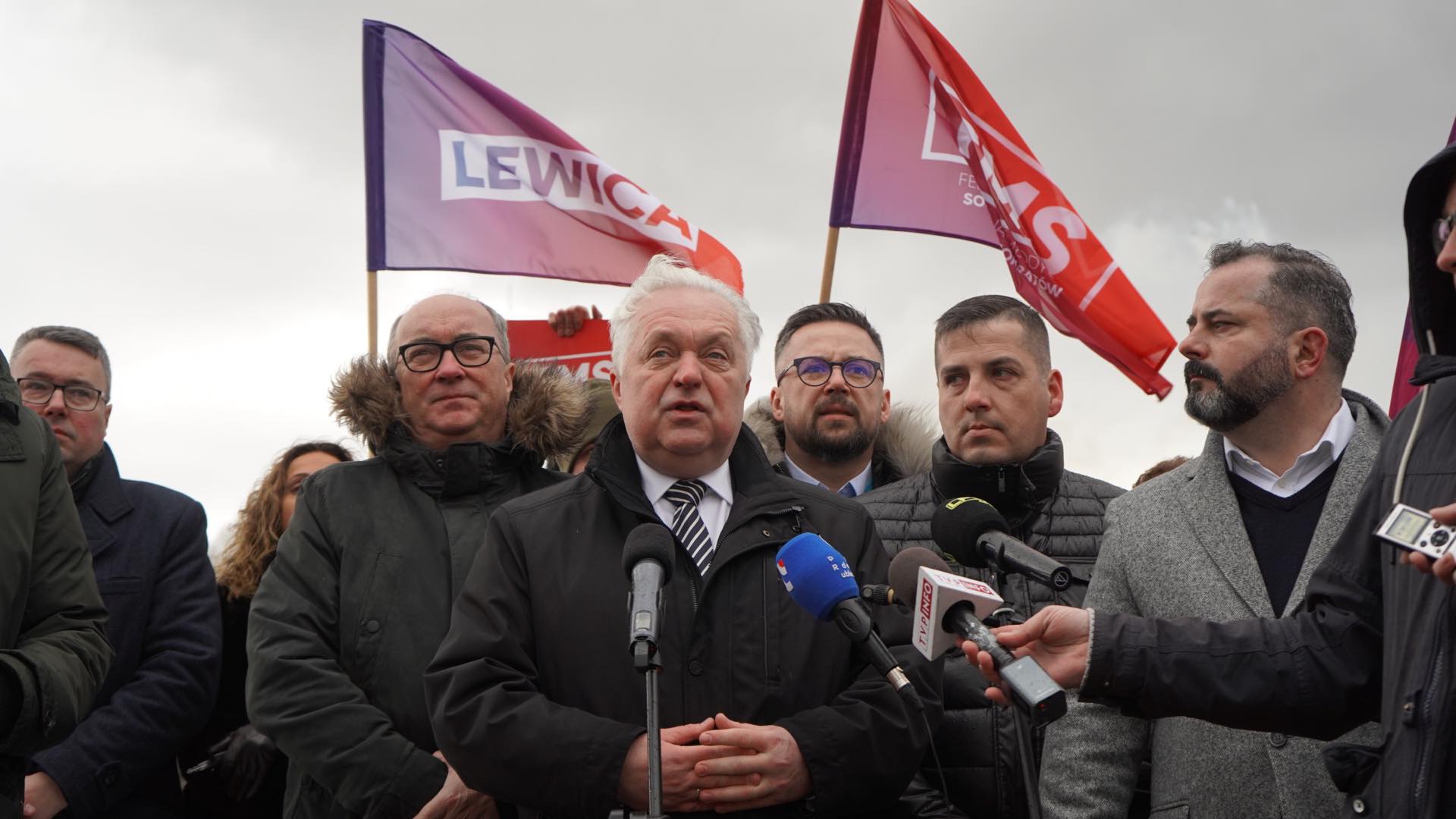 Konferencja prasowa przedstawicieli partii Lewica z flagami w tle.