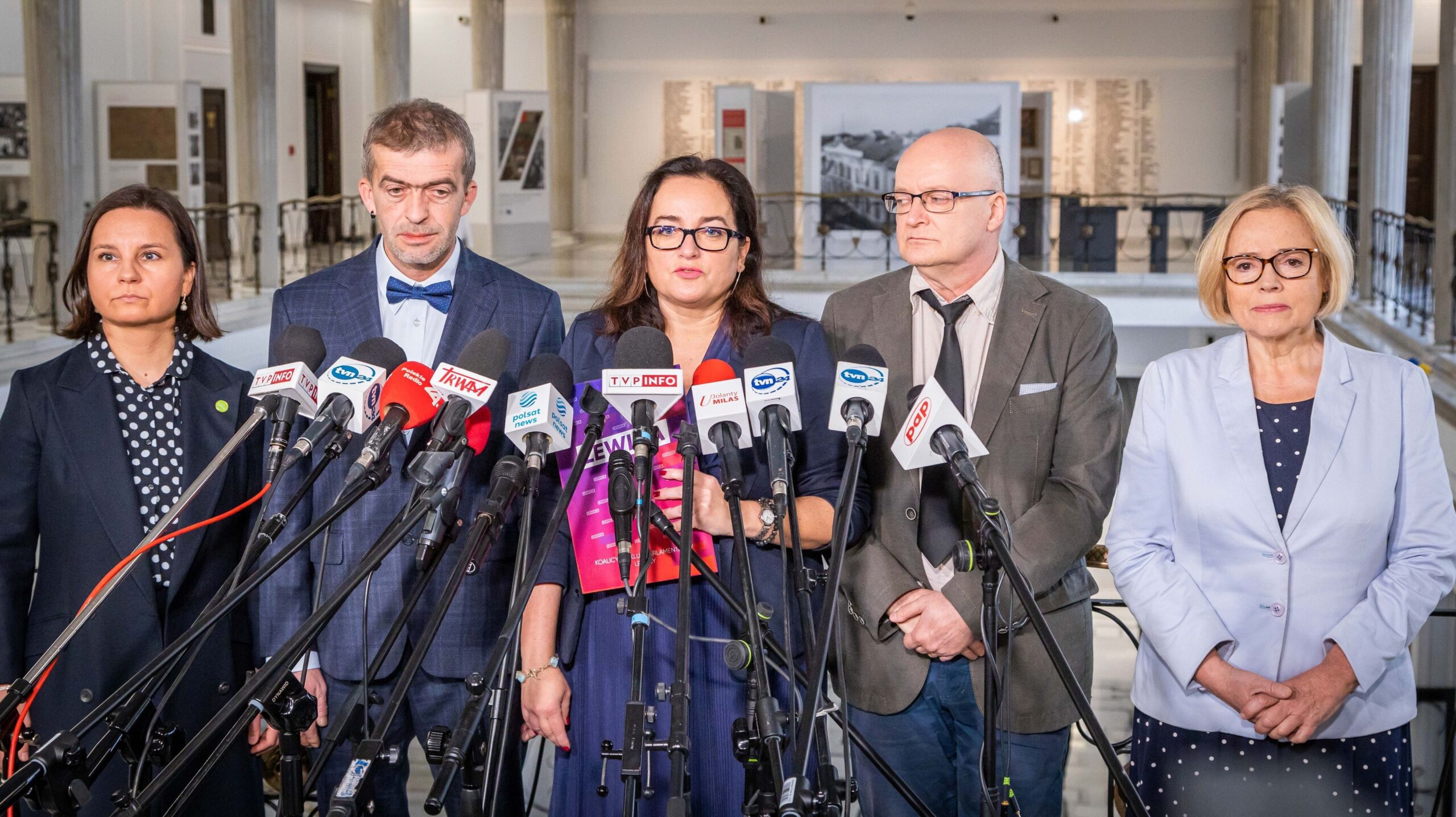 Na zdjęciu z prezentacją występuje pięć osób, które stoją przed mikrofonami, przygotowując się do konferencji prasowej w eleganckim wnętrzu.