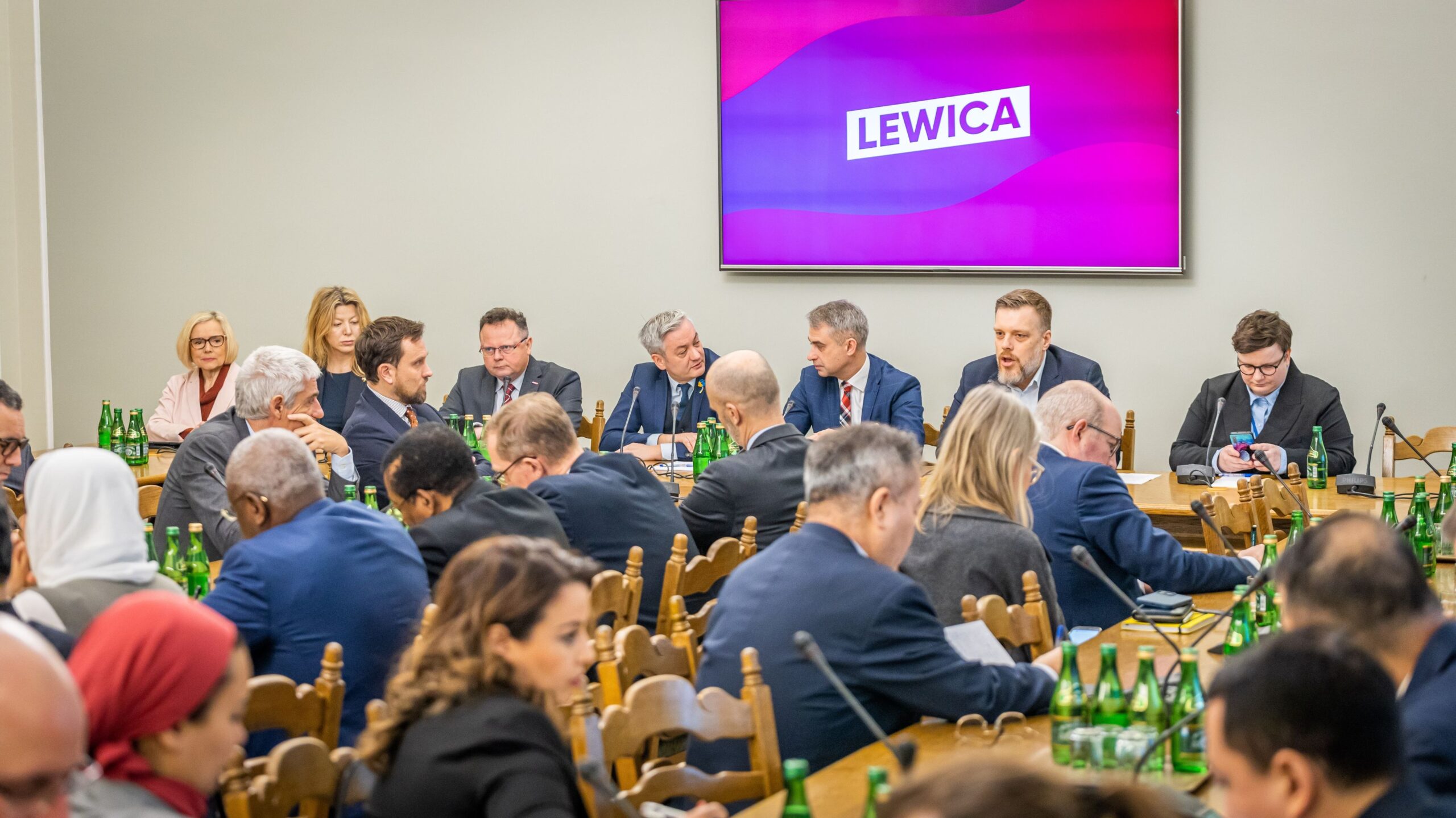 Na zdjęciu widać spotkanie polityków w sali konferencyjnej, przy stole z butelkami zielonej wody i ekranem z logo 