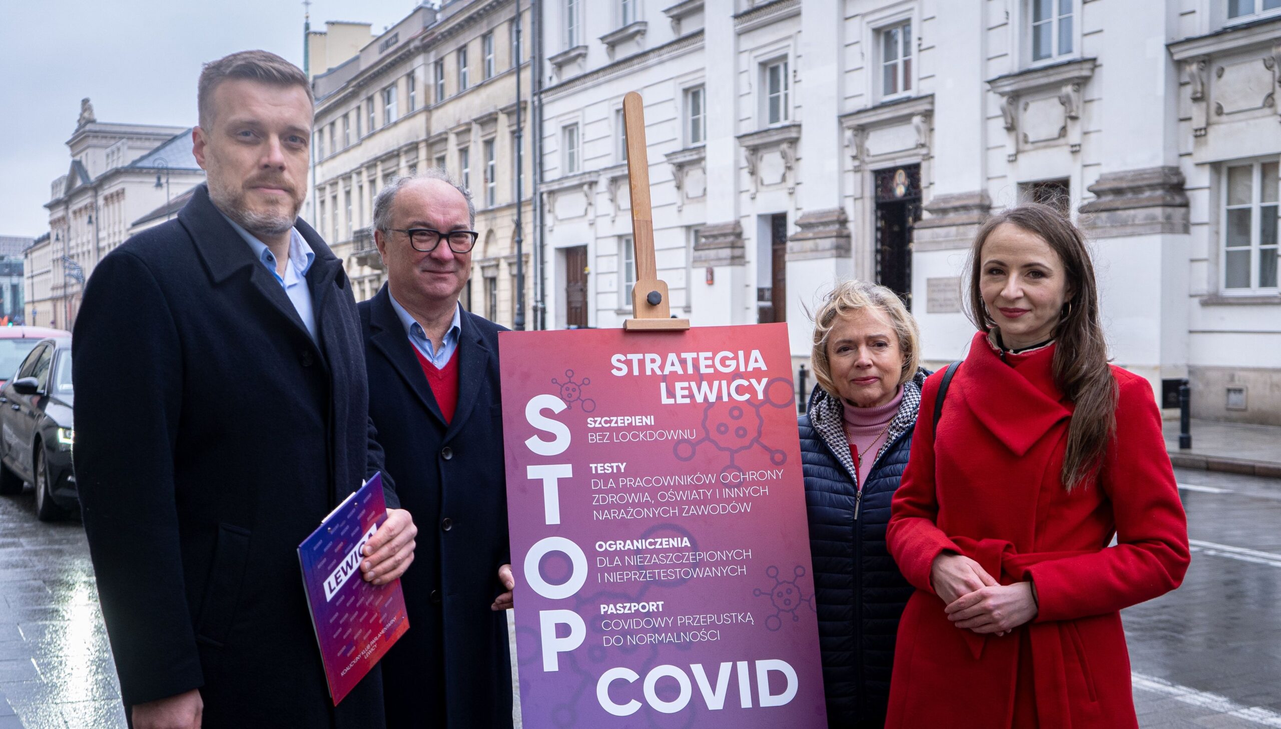 Czterech polityków stoi przed tablicą z hasłami dotyczącymi strategii Lewicy w kontekście COVID-19.